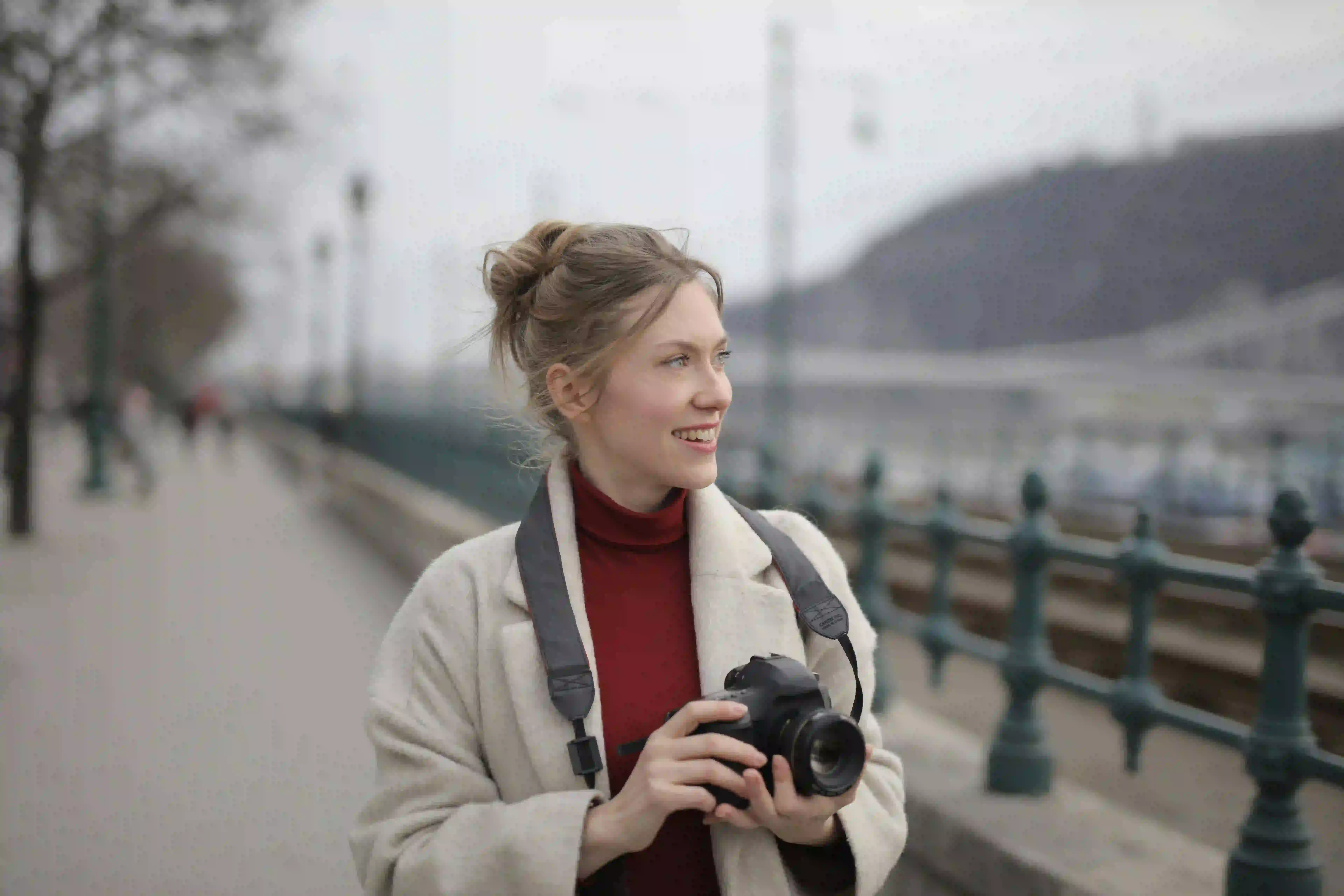 A smiling young woman in a cream coat and red turtleneck holds a camera while walking along a misty riverside. Her serene expression and observant gaze capture the essence of a mindful color walk in a calm, outdoor setting